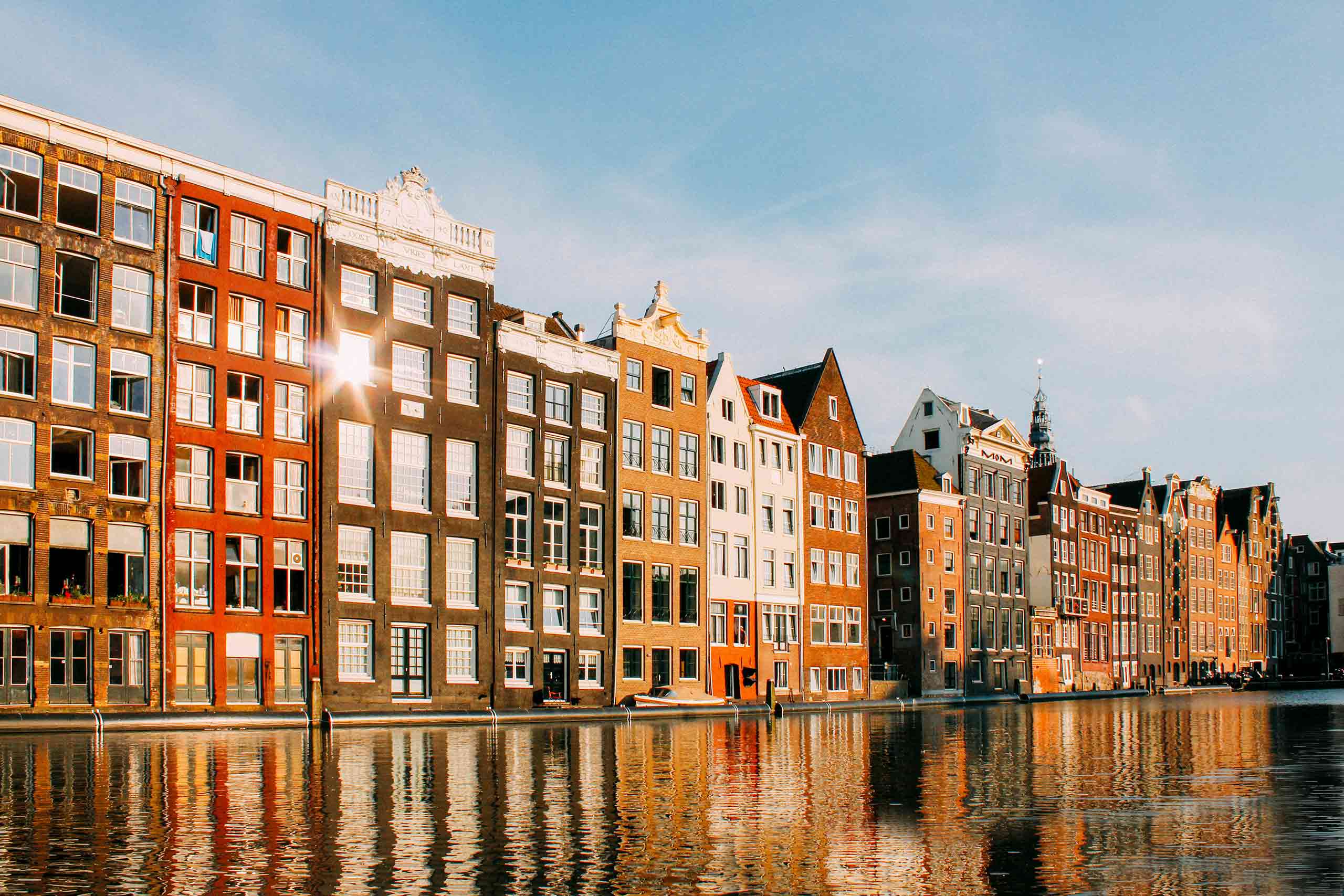 A row old Amsterdam canalside houses glow brown, red, orange and green in golden hour light. Behind them is one of the most OutThere hotels in Amsterdam