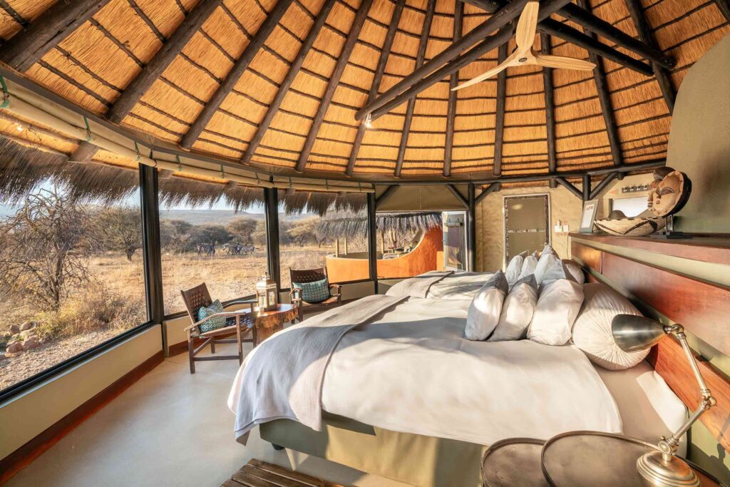 Two large beds dressed in white linen face a view of the African bush as zebras walk past at Okonjima Luxury Bush Camp, Okonjima Nature Reserve, Namibia