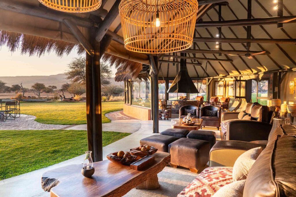 A comfortable seating area with wooden furniture and large lampshades looks out over the green plains at Okonjima Luxury Bush Camp, Okonjima Nature Reserve, Namibia