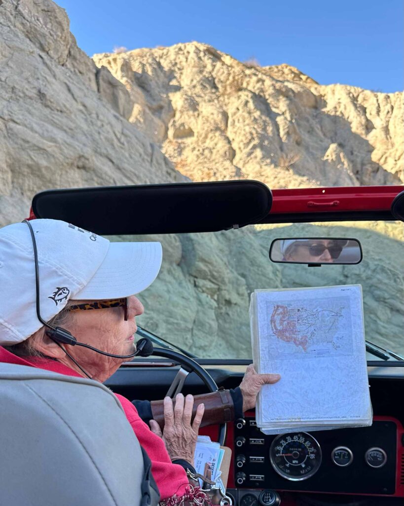 Red jeep tour guide Morgan sits in the drivers seat of a jeep holding a map and showing everyone sitting in the back. She's a white woman in her 70s and she wears a white baseball cap and headset