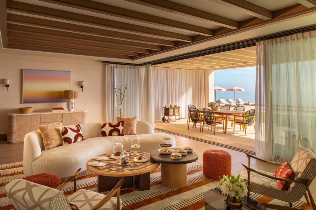 Welcome amenities await on two tables in front of a large sofa, with an opening at the back of the room giving guests a view of an outdoor dining area and poolside loungers beyond, Royal Mansour Tamuda Bay, Tamuda Bay, Morocco