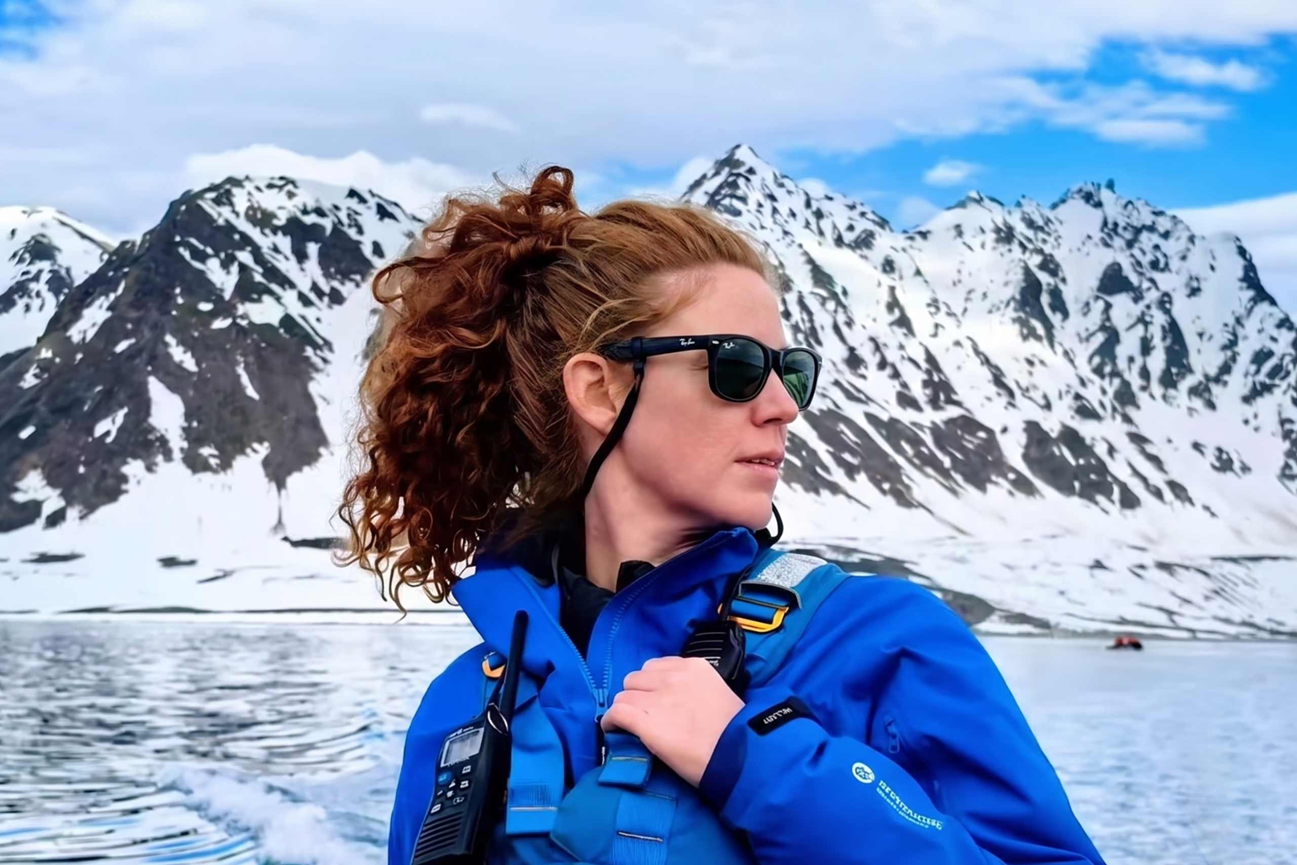 Portrait of Sarah Schwimmer, wearing ray ban sunglasses, a blue jacket and walkie-talkie in Svalbard. She is a white woman, with long red hair