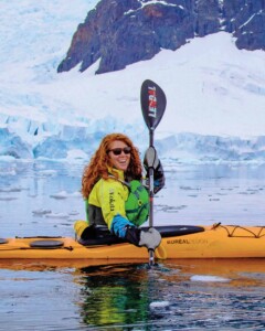 Sarah Schwimmer rows a yellow kayak in an arctic landscape, wearning a bright yellow jacket and sunglasses. She is a white woman, with long red hair and she is looking directly at the camera and smiling
