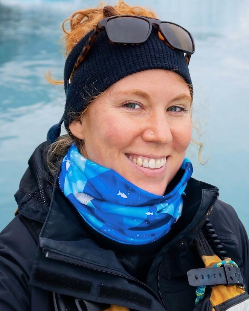 Portrait of Sarah Schwimmer, wearing a black hat with sunglasses on her head, a blue snood and a midnight blue jacket in Svalbard. She is a white woman, with long red hair and she is looking directly at the camera and smiling