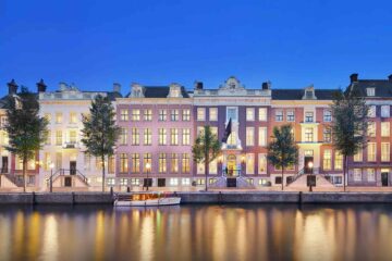 Six Amsterdam townhouses link together to form the Waldorf Astoria Amsterdam – this picture depicts the townhouses photographed at twilight, all in different dutch styles, from the other side of the canal, the lights of their windows reflecting in the water. In the midground, a white canal boat floats on the water