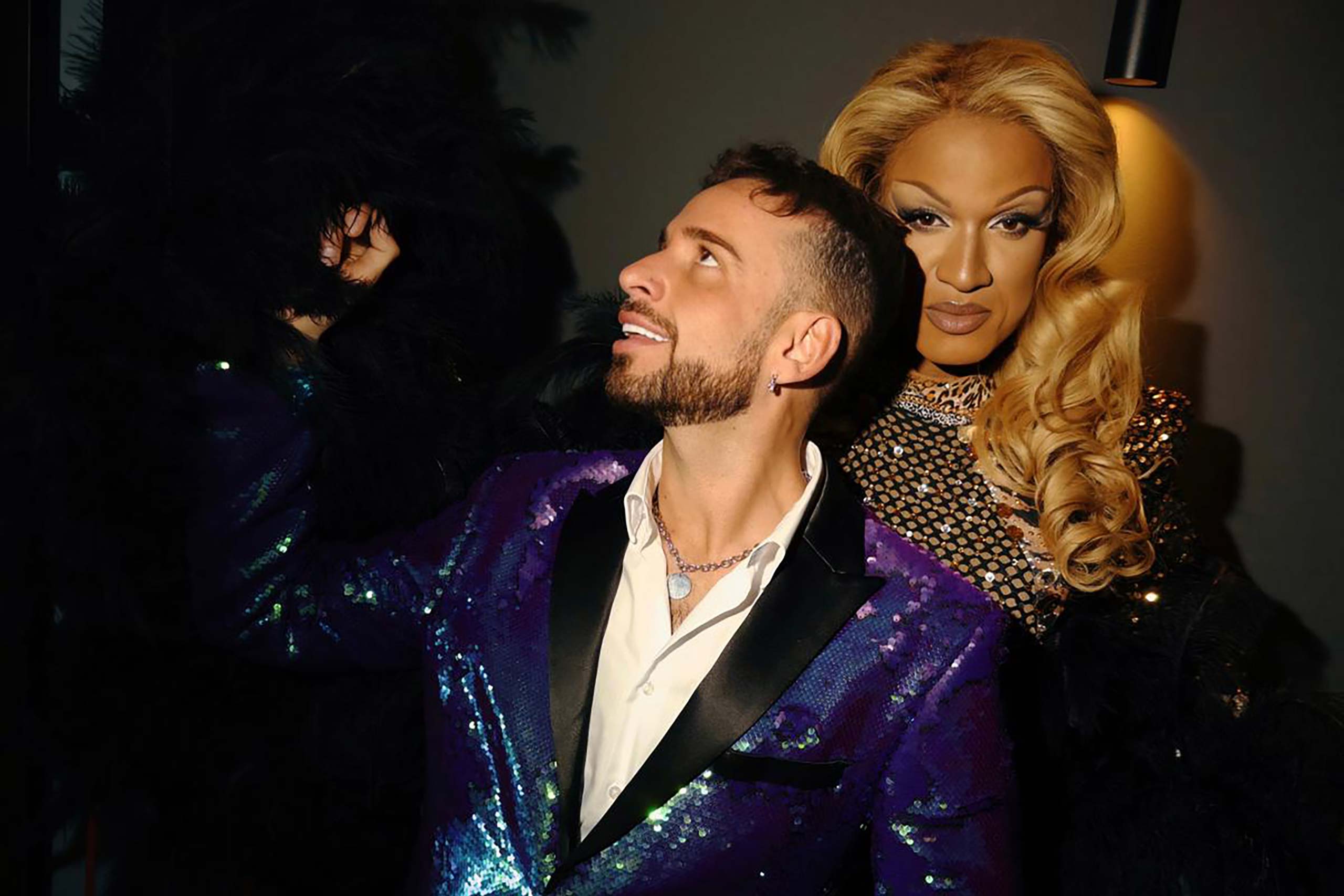 Amsterdam-based entertainer and content creator Gio Diekema stands in front of a drag queen. He has tanned skin, black hair and a moustache and beard. He wears a purple sequined tuxedo with black lapels and a white shirt unbuttoned to show his necklace and chest hair. The drag queen is of Afro-Caribbean descent and has beautiful curly blonde hair and a black and gold mesh dress. They are performing for WorldPride Amsterdam at Avani