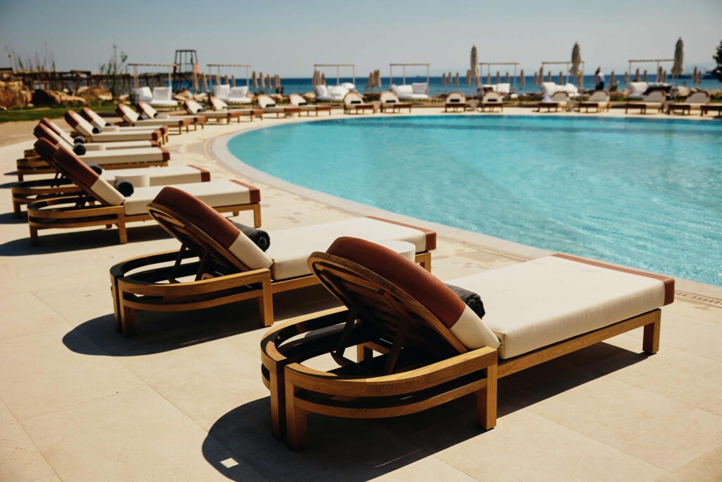 Poolside at 91 Athens Riviera, designer deck chairs with rounded top cusions flank a pool. In the background, the sea