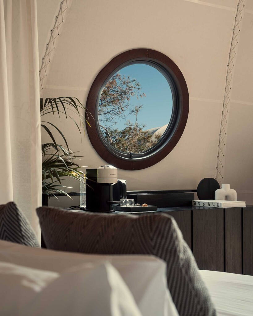 Tent interiors at 91 Athens Riviera, view from bed out of a porthole window. On a shelf is a Nespresso coffee maker, coffee table book and vases