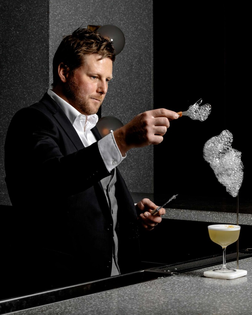 A mixologist wearing a black suit adds a cloud-like foam to a cocktail
