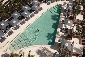 Aerial shot from the balcony of the Four Seasons Hotel and Residences Fort Lauderdale onto the pool deck, its central, curved – almost triangular shaped pool – flanked to the left and right by square, grey sun umbrellas and sunbeds. A woman with long brown hair swims in the pale blue water