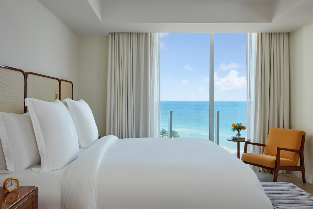Bedroom at the Four Seasons Hotel and Residences Fort Lauderdale, with pressed white sheets and fluffy pillows and brown wood furnishings – headboard, a side table with a copper alarm clock on it and an armchair with mustard upholstery. The room has a view of the sea through floor to ceiling windows