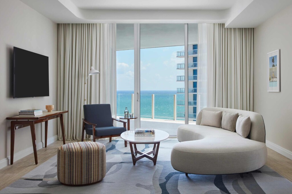 Living area at the Four Seasons Hotel and Residences Fort Lauderdale, with curved sofa, coffee table, striped ottoman and blue armchair with a white, mid-century style lamp over the top. On the wall a flat screen TV, and under it a console table with pottery and coffee table books. There are floor to ceiling sliding glass doors that lead out to the balcony that has a view of the ocean an to the balconies of the tower next door