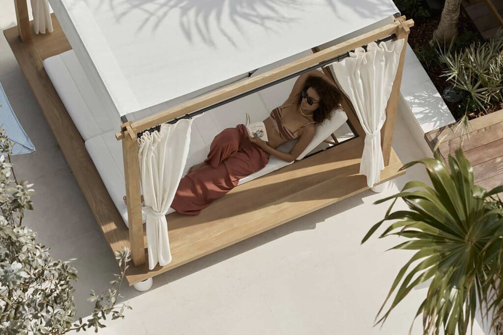 A woman relaxes with a book under a white and beech cabana. She wears a rust coloured loose trousers and a tripy brown bikini top, white necklace and sunglasses. The woman looks to be of African American origin and has big, curly brown hair