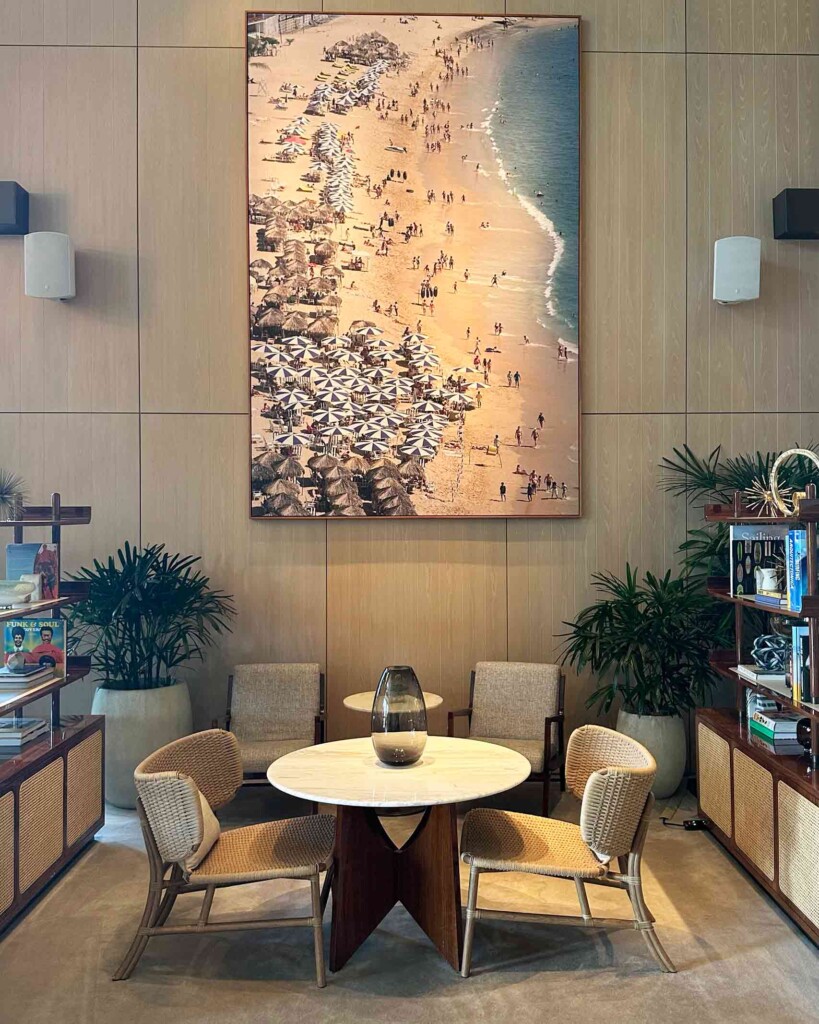 Lobby area at the Four Seasons Hotel and Residences Fort Lauderdale, featuring mid-century modern style, Floridian modern furnishings – table, chairs and shelving stocked with books and small collectables. On the wall in the background is a huge photograph of Nik Wheeler’s, A La Playa