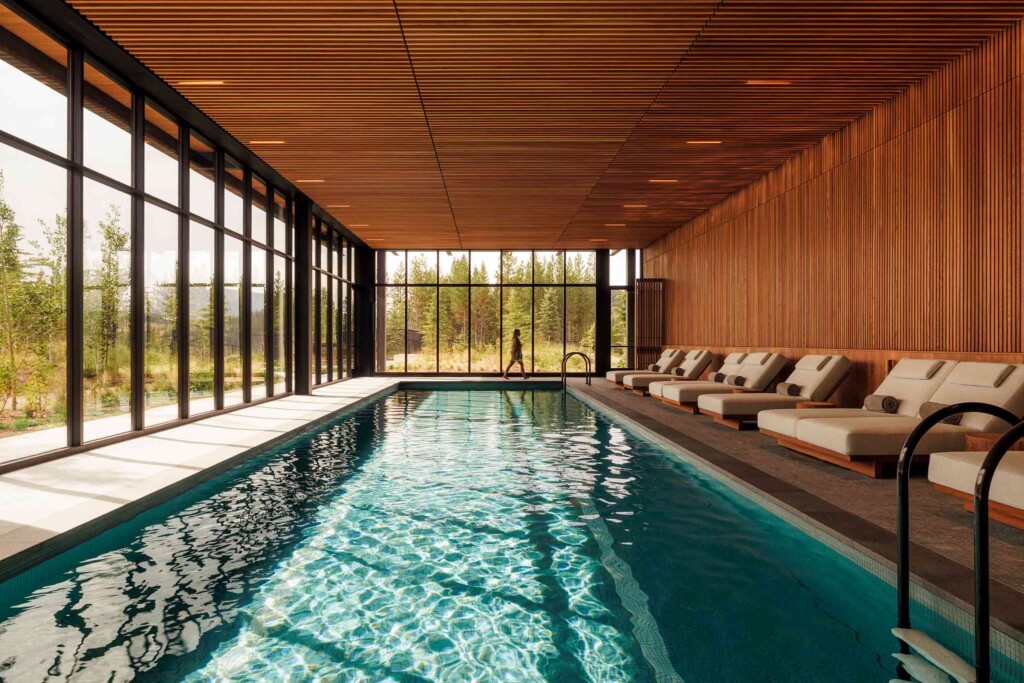 A guest walks towards padded loungers lined up next to a large indoor pool with views of the surrounding forests at One&Only Moonlight Basin, Big Sky, Montana, USA
