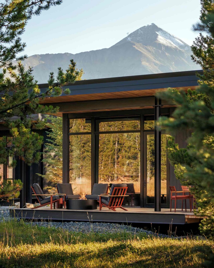 Low-slung armchairs surround a log fire in front of a glass-fronted private accommodation in front of a mountain in Big Sky, Montana