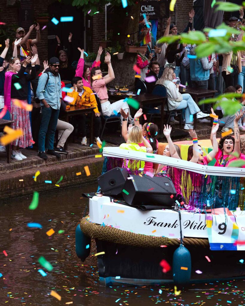 Confetti in the air as people party on a boat and a crowd watches from the adjoining canal side