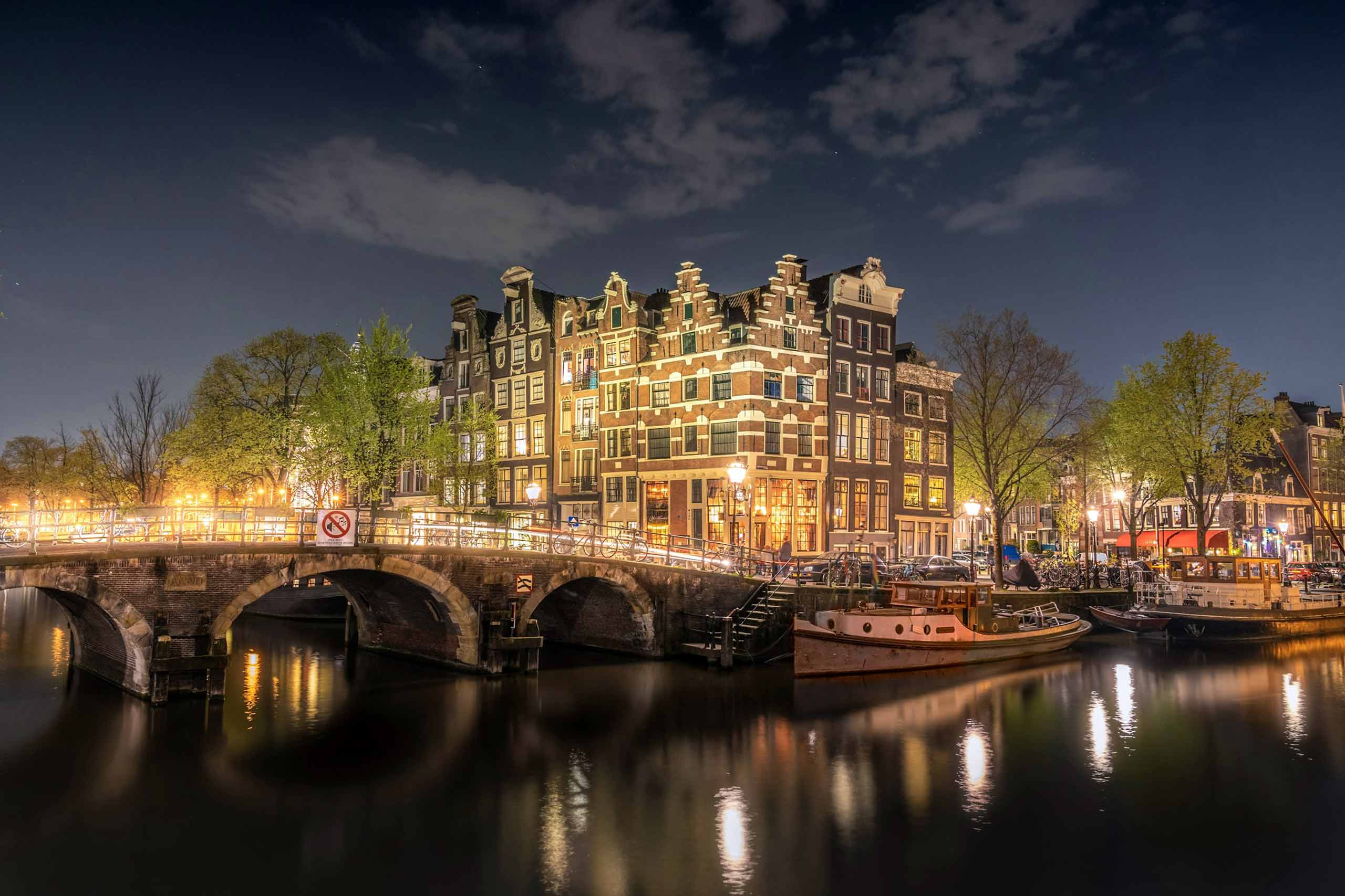 A bridge and a row of traditional Dutch houses are brightly illuminated at night in Amsterdam, an illustration of the upcoming WorldPride Amsterdam 2026