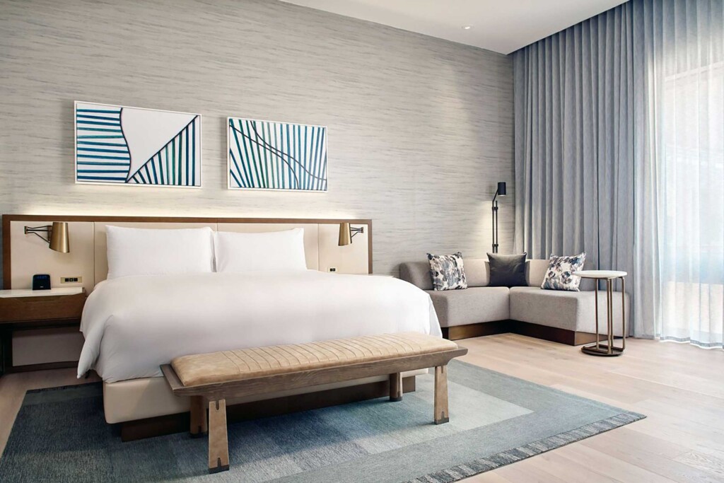 A king-size bed with a large headboard and a corner bench sit alongside one another in a beige and light blue Junior Suite at JW Marriott Hotel Mexico City Polanco, Mexico City, Mexico