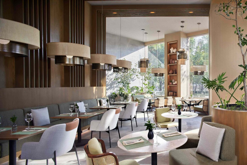 Large lampshades hang above rows of tables set for lunch in front of large windows at Sendero Restaurant in CDMX