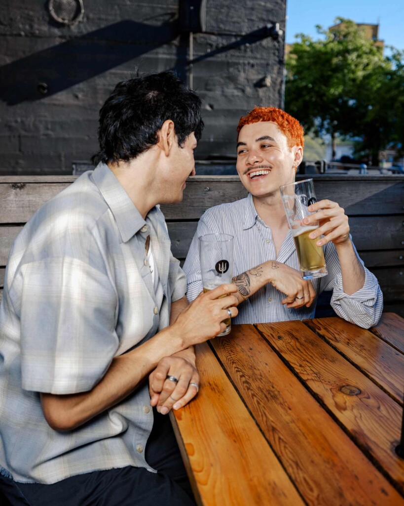 Two young men sit at a table in a Portland pub, laughing and drinking beer together, an example showcasing the open-minded mentality of the city Jenny Nguyen has built her career on