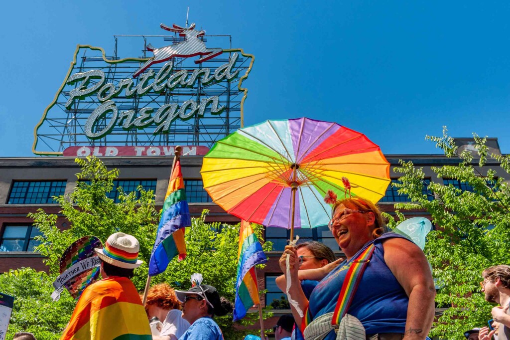 During a Pride march, event attendees dressed in rainbow flags and holding a rainbow flag umbrella, march outside buildings in Portland, Oregon, an example showcasing the open-minded mentality of the city Jenny Nguyen has built her career on