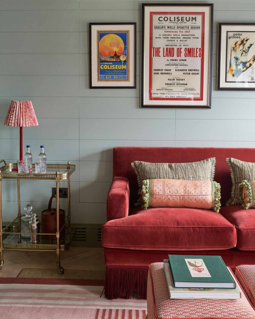 A cosy sitting area at Beaverbrook Town House with a deep rose-red velvet sofa, patterned cushions, a brass drinks trolley, and vintage theatre posters on soft grey panelled walls, creating a refined, theatrical atmosphere.