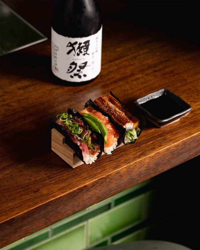 Three hand rolls filled with different seafood sit on a wooden counter beside soy sauce, with a bottle of Japanese sake in the background under warm, moody lighting.