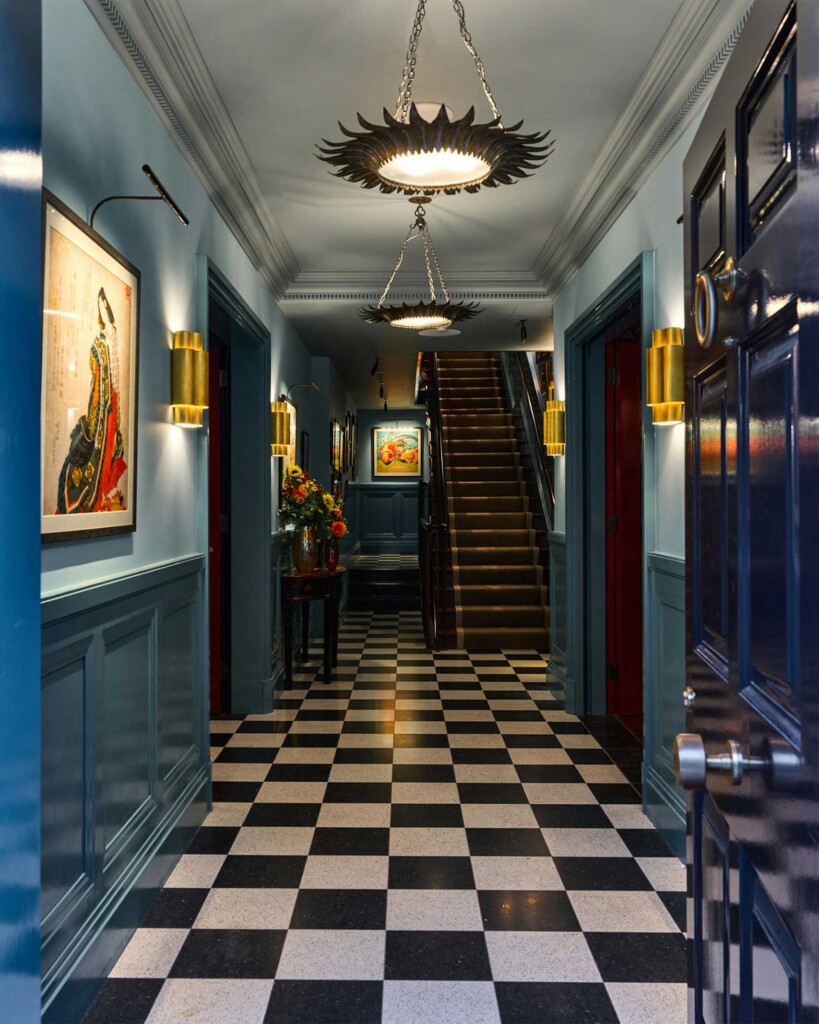 A long hallway at Beaverbrook Town House with black-and-white chequerboard flooring, blue panelled walls, striking brass light fittings, colourful artwork, and a staircase leading to the upper floors.