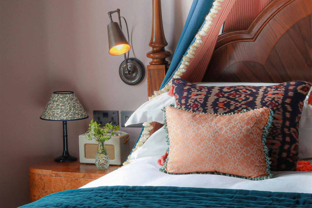 A carved wooden four-poster bed dressed in crisp white linens and colourful patterned cushions sits beside a vintage-style lamp, Roberts radio, and fresh flowers against soft pink walls.