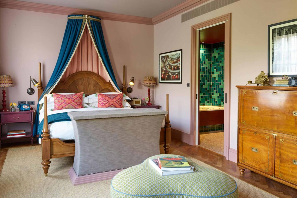 An elegant bedroom at Beaverbrook Town House featuring a wooden four-poster bed with dramatic blue canopy curtains, pastel pink walls, antique furniture, and a green-tiled bathroom visible through an open doorway.