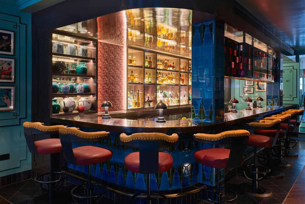 A richly coloured cocktail bar with glossy blue tiles, illuminated shelves of spirits and ceramics, polished metal surfaces, and leather bar stools arranged around a central counter.
