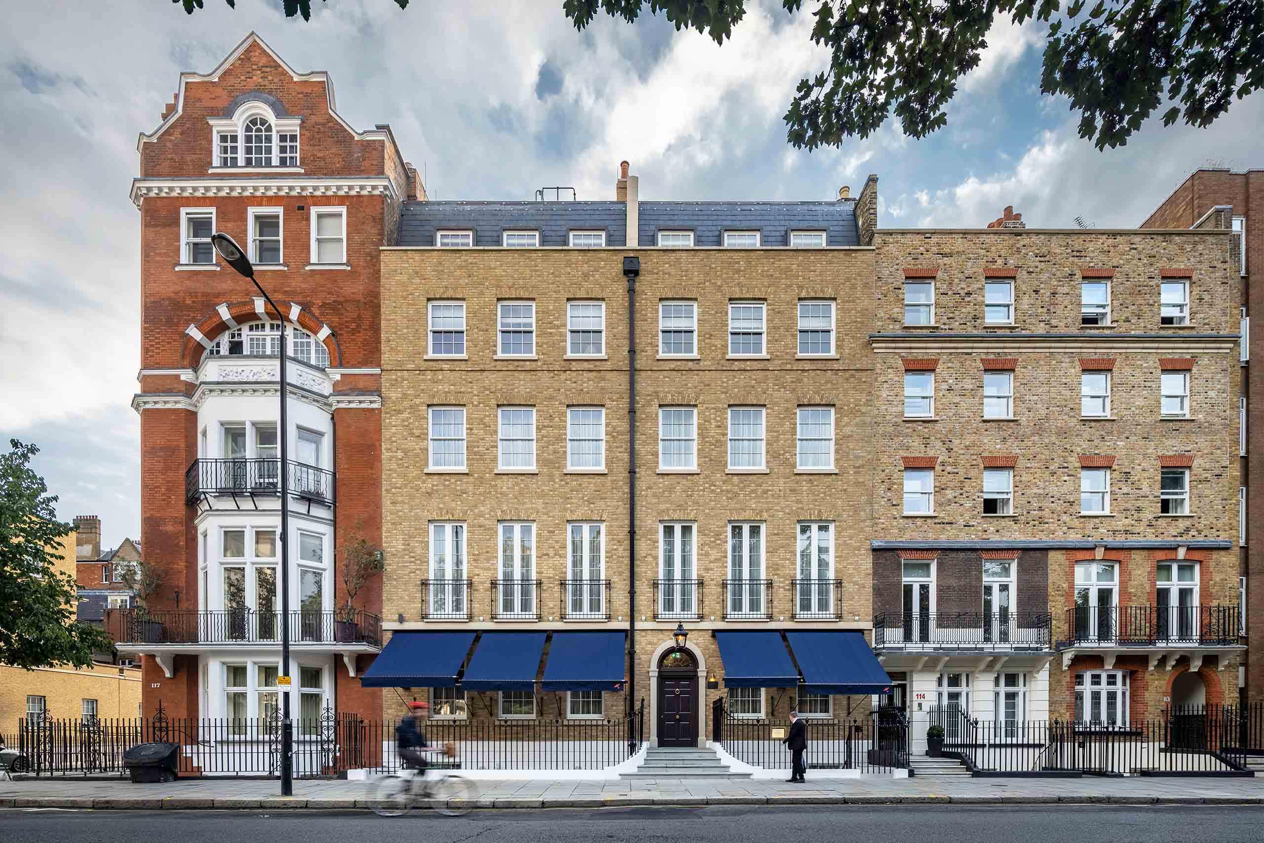 The hotel's elegant Georgian townhouse façade features honey-coloured brickwork, navy awnings, tall sash windows, and a central entrance overlooking a quiet Chelsea street.