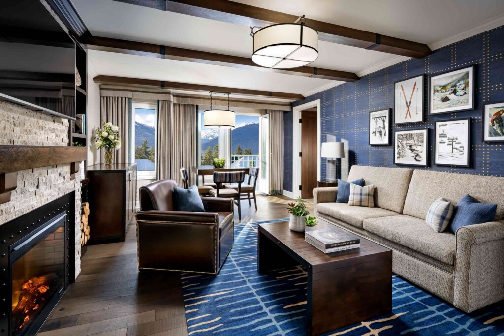 A large TV hangs above a fireplace in a lounge space dressed in blue rugs, deep armchairs and wintersports-related wallart, inside a suite at Fairmont Chateau Whistler, Whistler, Canada