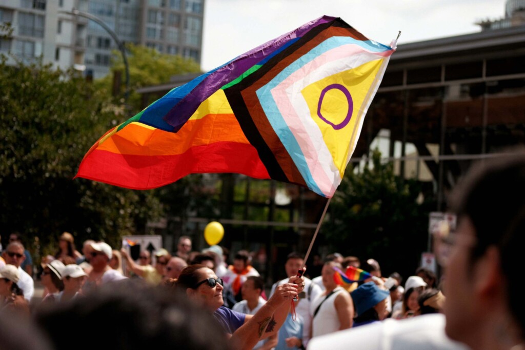 A woman waves a Progress flag amid a large crowd of Pride attendees, a symbol of an Intersectional Amsterdam