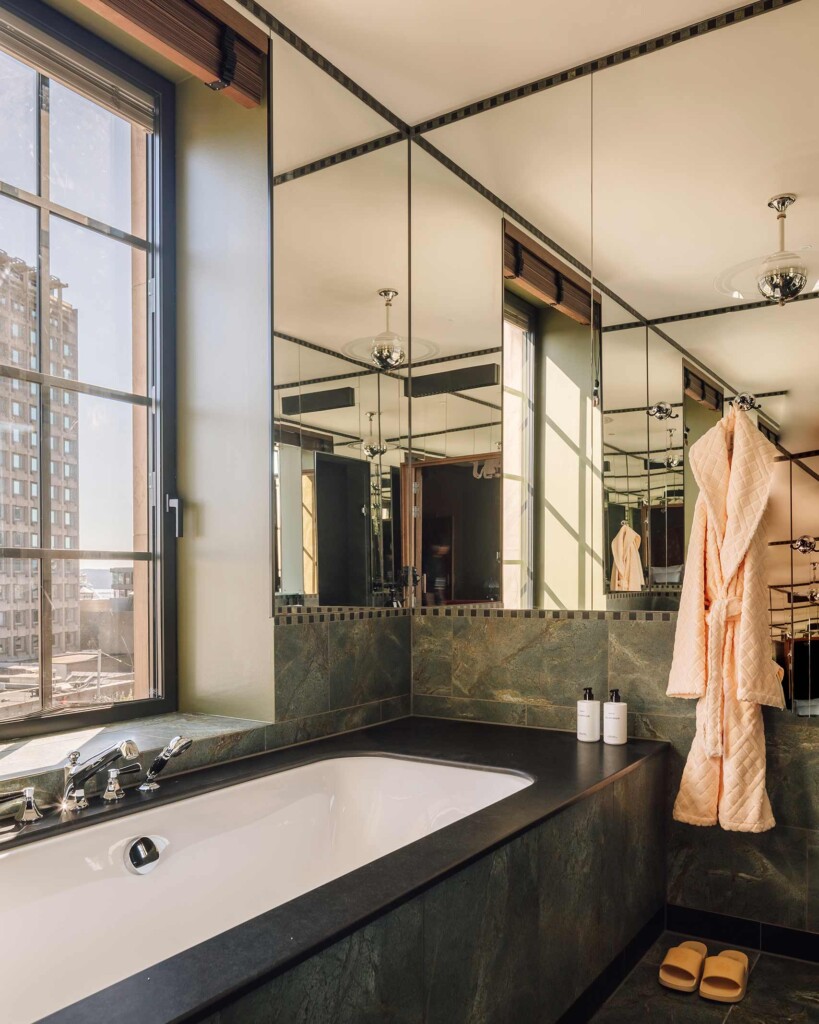 A large built-in bathtub in a dark green marble bathroom stands by a large window and is surrounded by mirrored walls, Sommerro, Oslo, Norway