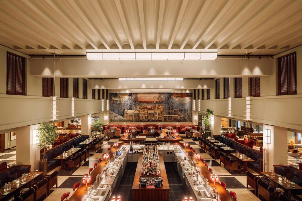Art Deco ceiling lights hang above a large, open-air space within which a restaurant and bar sit with a large, industrial mural on the back wall, Ekspedisjonshallen, Oslo, Norway
